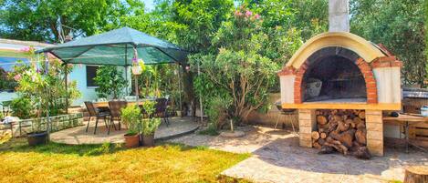 An outdoor patio area with a brick pizza oven, a covered seating area with chairs, and a stone pathway.