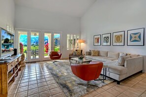 A spacious living room with a large TV, a red chair, a beige sofa, and a coffee table. There are framed pictures on the wall and a view of a garden through the sliding doors. - Santa Rosa Cove #50, 3 Bedrooms 3.5 Baths La Quinta City Permit 063498 (La Quinta)