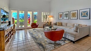 A spacious living room with a large TV, a red chair, a beige sofa, and a coffee table. There are framed pictures on the wall and a view of a garden through the sliding doors. - Santa Rosa Cove #50, 3 Bedrooms   3.5 Baths  La Quinta City Permit 063498 (La Quinta)