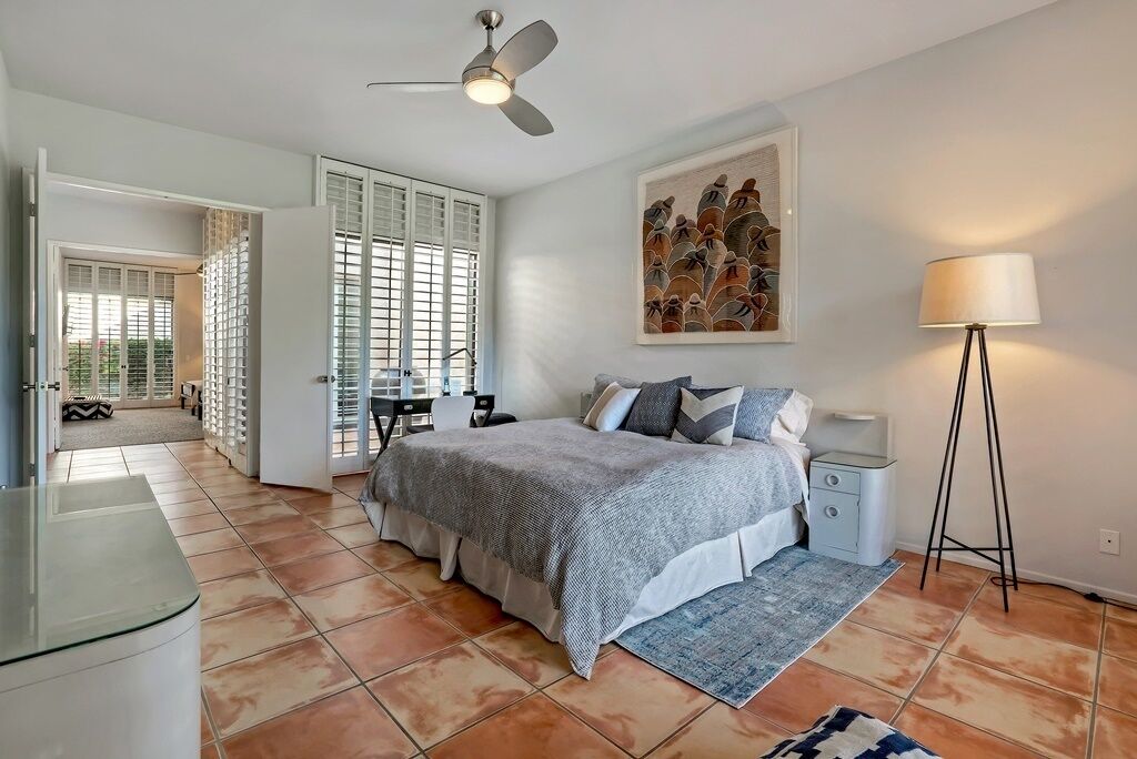 Um quarto com cama, ventoinha no teto, mesa de cabeceira, abajur e um quadro na parede.