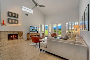 A spacious living room with a fireplace, a ceiling fan, a TV, and a large window with a view of the outdoors. - Santa Rosa Cove #50, 3 Bedrooms 3.5 Baths La Quinta City Permit 063498 (La Quinta)