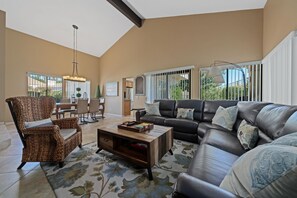 A spacious living room with a leather sofa, wicker armchair, and a coffee table. There is a dining area with a table and chairs, and a kitchen in the background. - Santa Rosa Cove #51, 3 BD/2 BA, Pet Friendly Home, Permit 063499 (La Quinta)