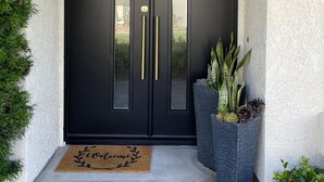 Eine schwarze Doppeltür mit goldenen Griffen, eine Fußmatte mit dem Schriftzug 'welcome' und ein Pflanzgefäß mit Pflanzen neben der Tür.