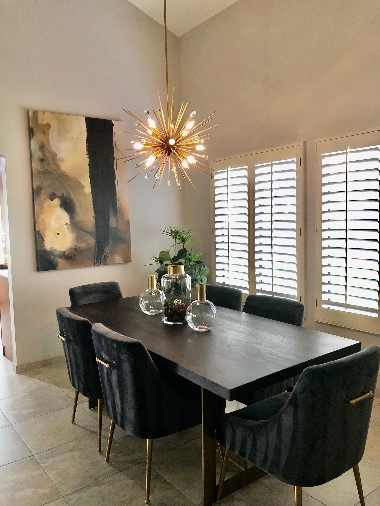 A modern dining room with a rectangular table, dark chairs, a unique chandelier, and a large abstract painting on the wall.