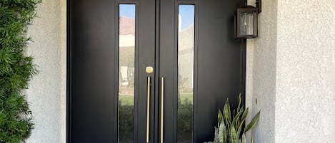 A black double door with gold handles, a doormat with 'welcome' written on it, and a planter with plants beside the door.