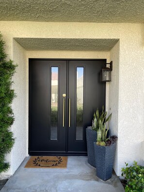 A black double door with gold handles, a doormat with 'welcome' written on it, and a planter with plants beside the door. - Santa Rosa Cove #57, 3 bedroom/2bath, Permit 063491 (La Quinta)