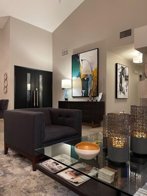 A modern living room with a dark sofa, a glass coffee table, magazines, decorative items, and abstract paintings on the walls. - Santa Rosa Cove #57, 3 bedroom/2bath, Permit 063491 (La Quinta)