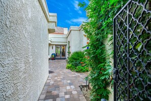 A narrow pathway leading to a building with a door, surrounded by greenery and a metal gate. - Beautiful Home on Golf Course with Amazing Mtn Views! Permit 063495 (La Quinta)