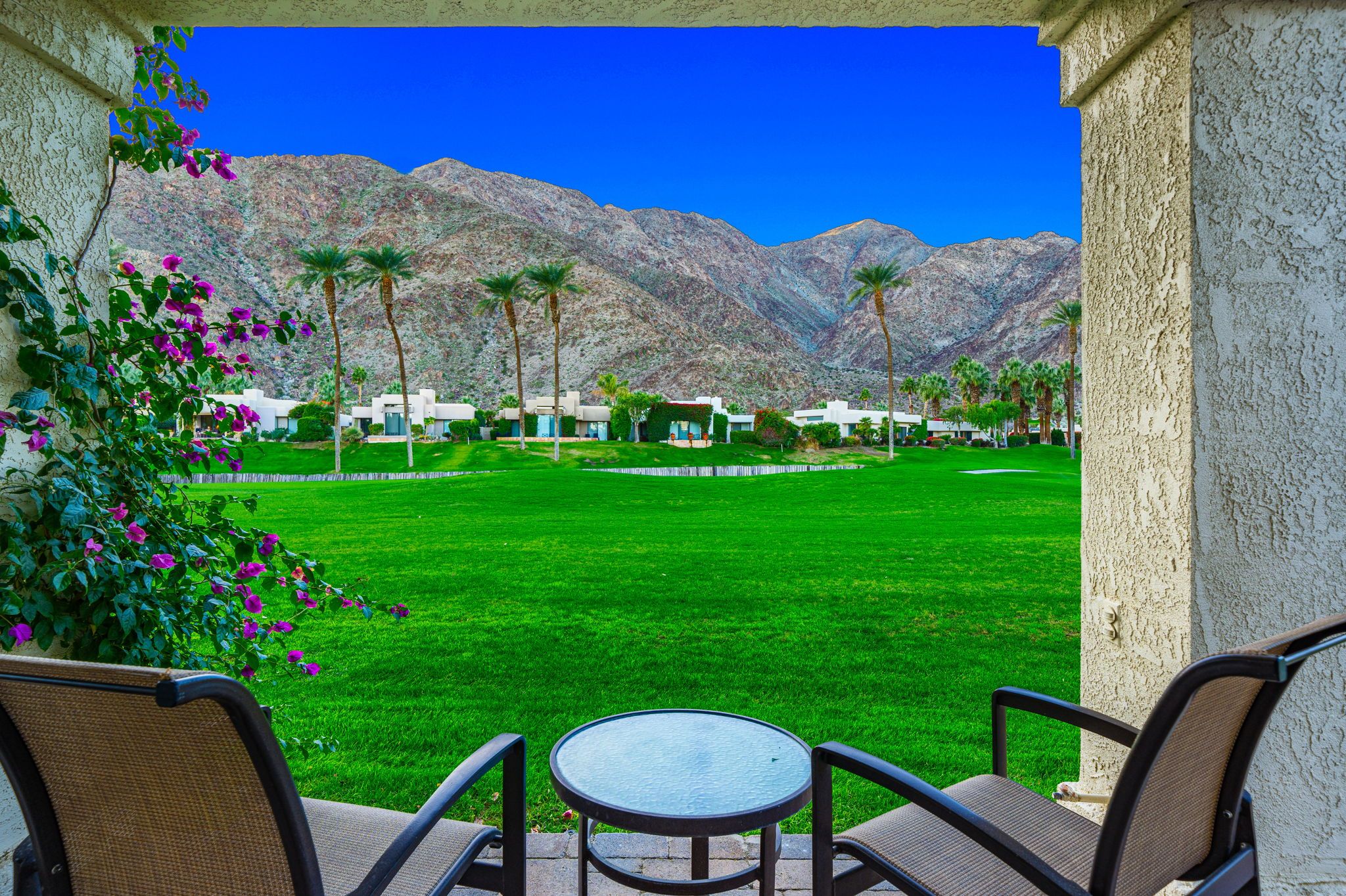 A patio with a table and chairs, overlooking a well-manicured lawn and a golf course, with mountains in the background.