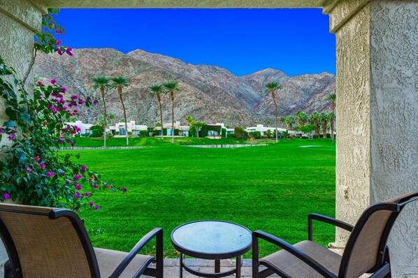 A patio with a table and chairs, overlooking a well-manicured lawn and a golf course, with mountains in the background. - Beautiful Home on Golf Course with Amazing Mtn Views! Permit 063495 (La Quinta)