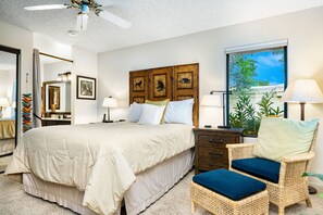 A bedroom with a large bed, a wicker chair with cushions, a nightstand, a window with a view of greenery, and a ceiling fan. - Beautiful Home on Golf Course with Amazing Mtn Views! Permit 063495 (La Quinta)