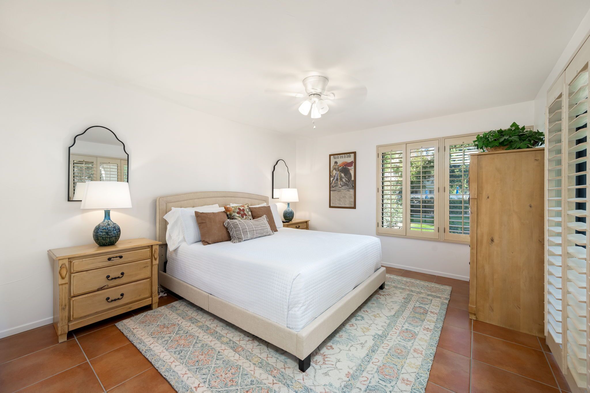A bedroom with a bed, bedside tables, a ceiling fan, a window with blinds, and a wooden cabinet.