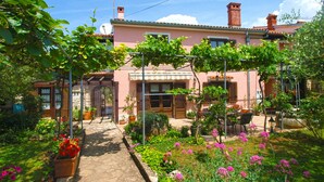 Una villa rosa con un camino de piedra que lleva a una puerta de madera, rodeada de exuberantes jardines y mobiliario de exterior.