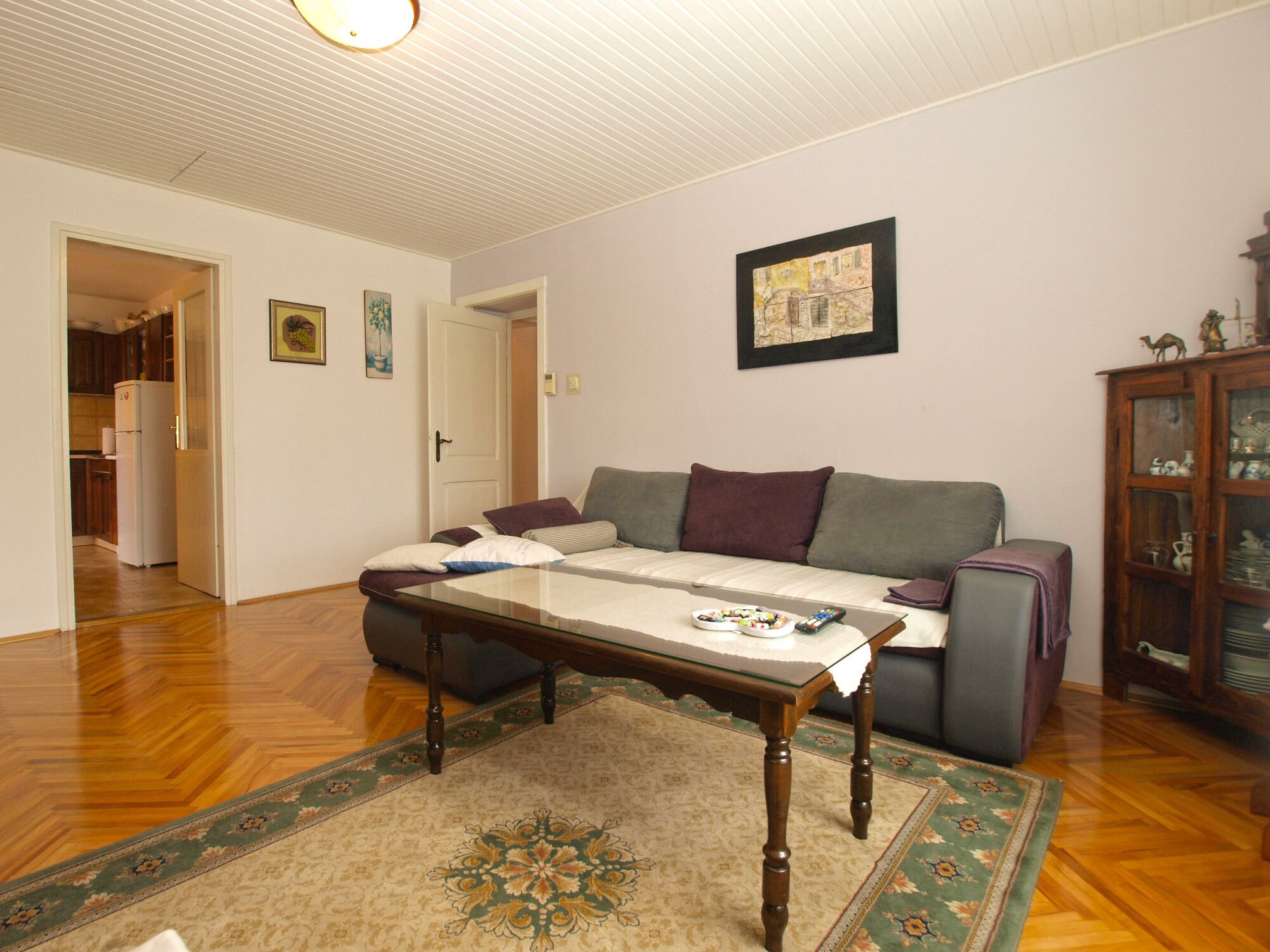 A living room with a sofa, a glass-top coffee table, a patterned rug, a wooden cabinet with decorative items, and a door leading to a kitchen.
