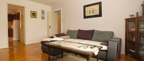 A living room with a sofa, a glass-top coffee table, a patterned rug, a wooden cabinet with decorative items, and a door leading to a kitchen.