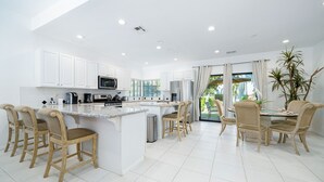 A modern kitchen with white cabinetry, a central island, and a dining area with wicker chairs. - Tennis Villa #10, Close to Festivals!   Pet Friendly Home.  Permit 063443 (La Quinta)