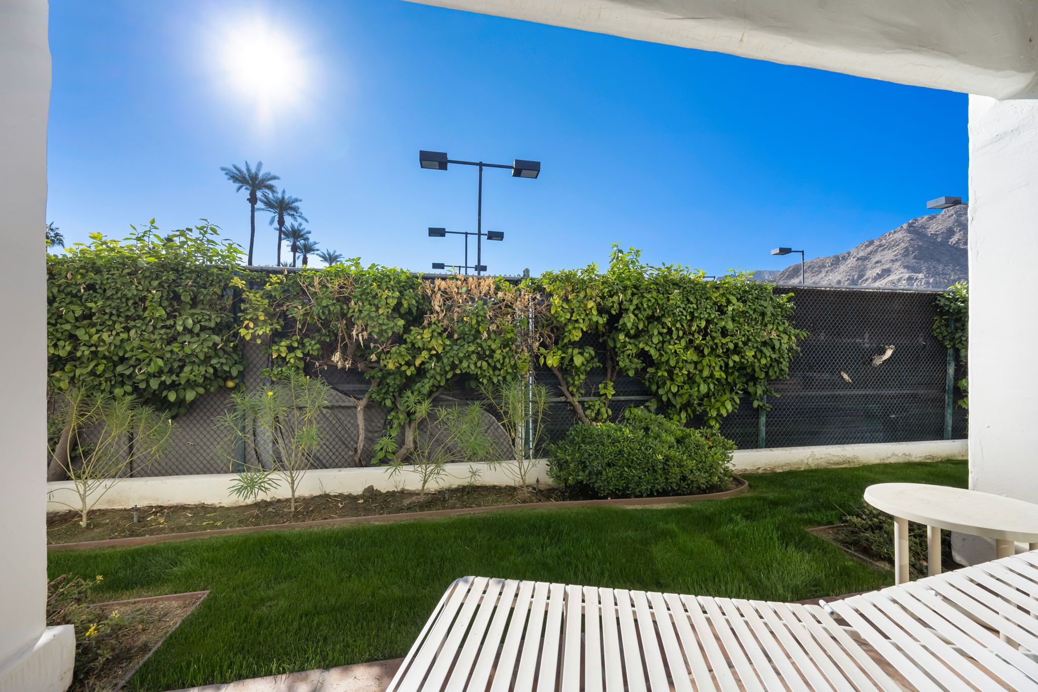 A patio area with a white table and chairs, a small garden with green grass and plants, and a view of a mountain and street lamps in the background.