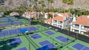 Un complejo de canchas de tenis con varias canchas, rodeado de edificios residenciales con techos de tejas rojas y palmeras.