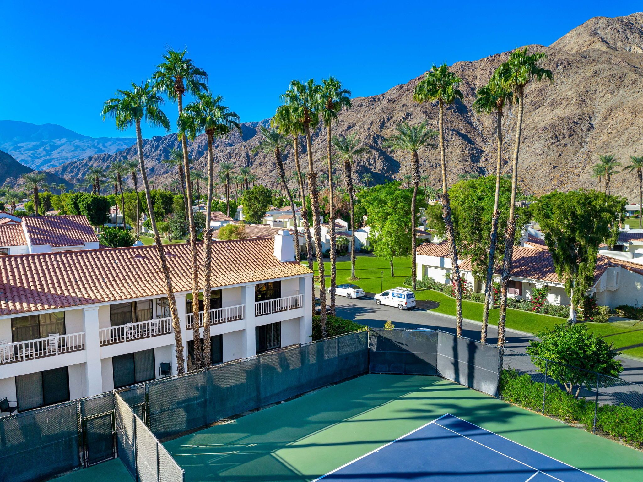 A residential area with a tennis court, white buildings with red-tiled roofs, and palm trees.