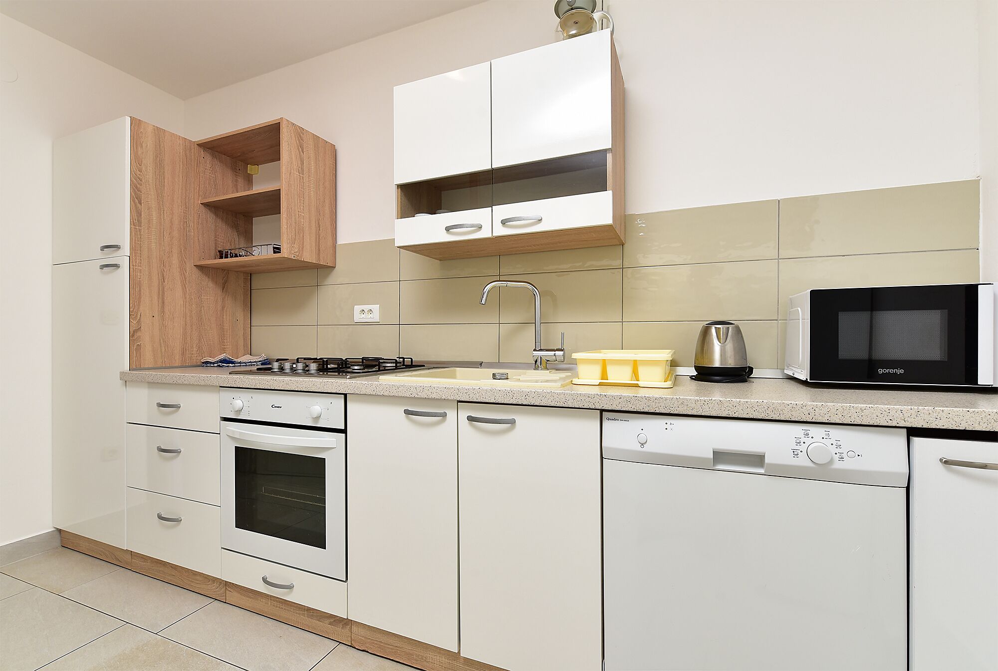A kitchen with white cabinets, a built-in oven, a dishwasher, a microwave, and a wooden shelf.