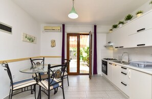 A compact kitchen with a dining area, a small table, chairs, a hanging light fixture, and a view through a door to an outdoor area. - Apartment 85/144 (Istria - Fažana), 50m from the beach (Fažana)