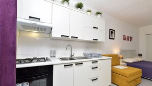 Eine kompakte Küche mit Herd, Spüle und Schränken, ein kleiner Essbereich mit Tisch und Stühlen sowie ein Schlafzimmer mit Bett und Nachttisch.