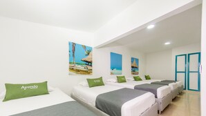A hotel room with four beds arranged in a row, each with a white mattress and grey bedding. There are green pillows with the hotel name on them. The walls are white, and there are two beach-themed paintings above the beds.