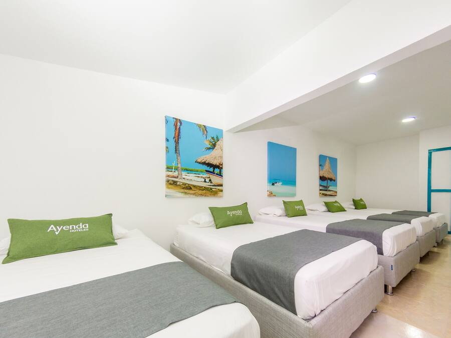 Quarto de hotel com quatro camas dispostas em linha, cada uma com colchão branco e roupa de cama cinzenta. Há almofadas verdes com o nome do hotel. As paredes são brancas e há duas pinturas com tema praiano acima das camas.