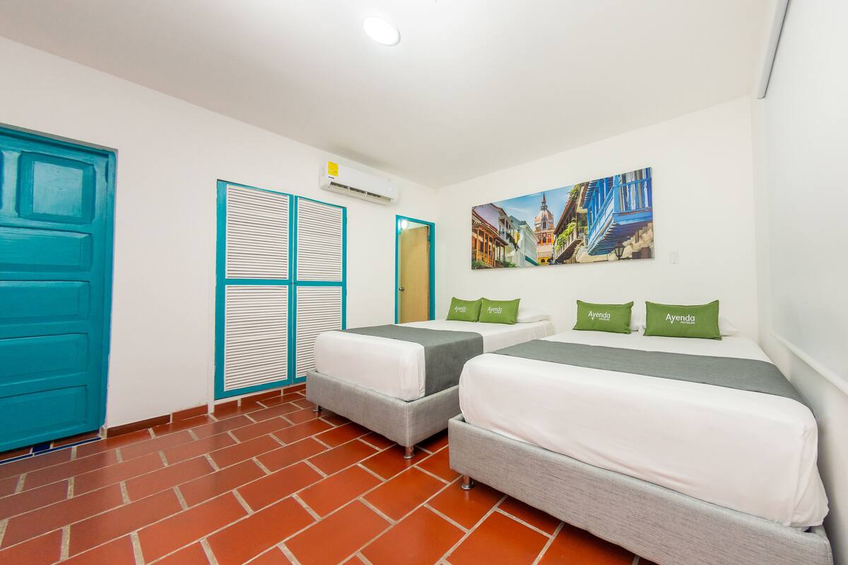 A hotel room with two beds, a blue door, a window with blinds, and a painting on the wall.