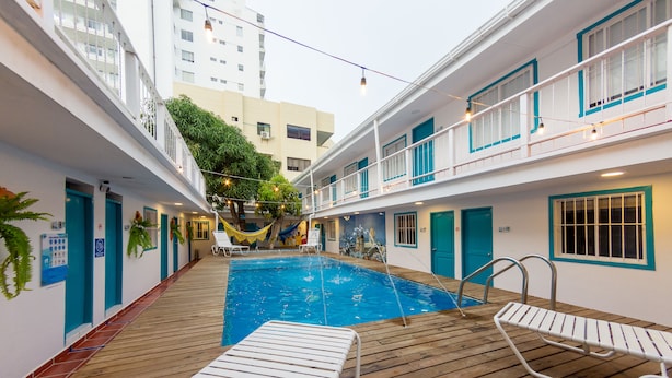 A pool area with wooden deck and lounge chairs, surrounded by a building with multiple balconies and windows.