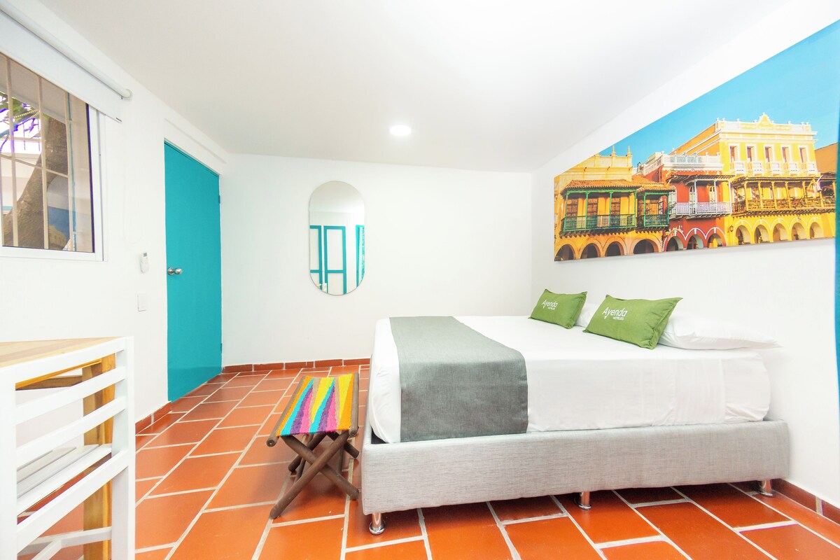 Une chambre d’hôtel avec un lit, un tabouret coloré, une fenêtre et une fresque représentant un bâtiment.
