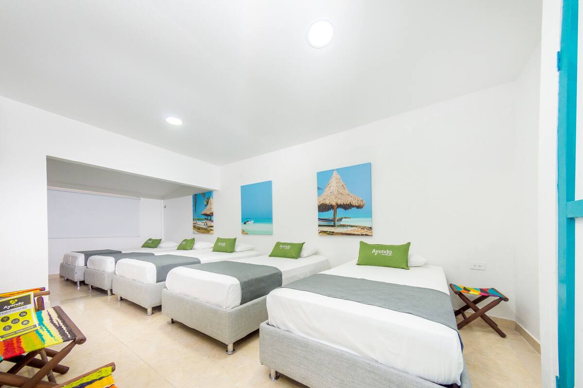 A hotel room with four single beds, each with a green pillow, white bedding, and a small wooden side table.