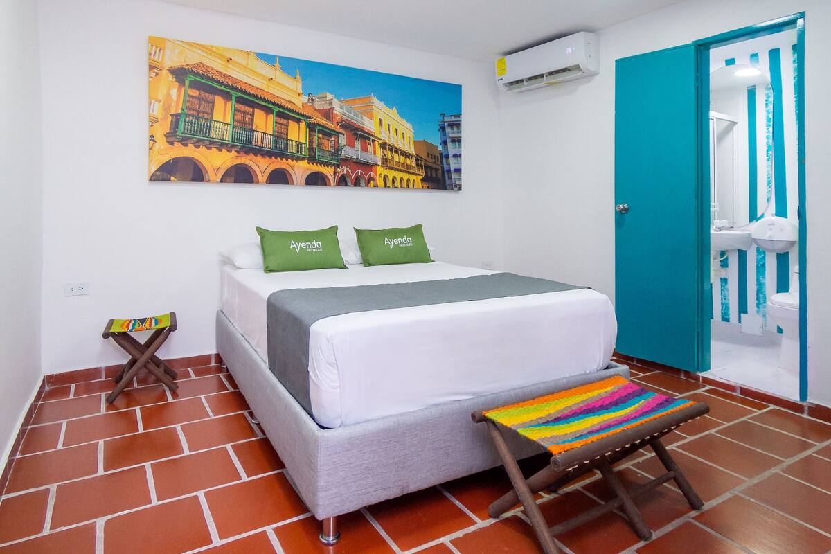 A hotel room with a bed, a colorful stool, a painting of buildings, and a blue door.