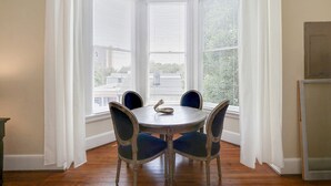 Un área de comedor con una mesa redonda y cuatro sillas, ventanas amplias con persianas y vistas a edificios.