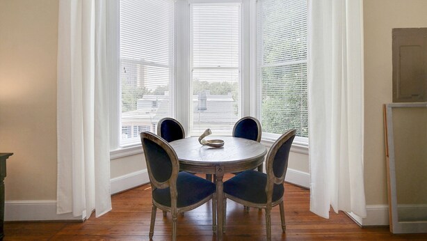 A dining area with a round table and four chairs, large windows with blinds, and a view of buildings outside.
