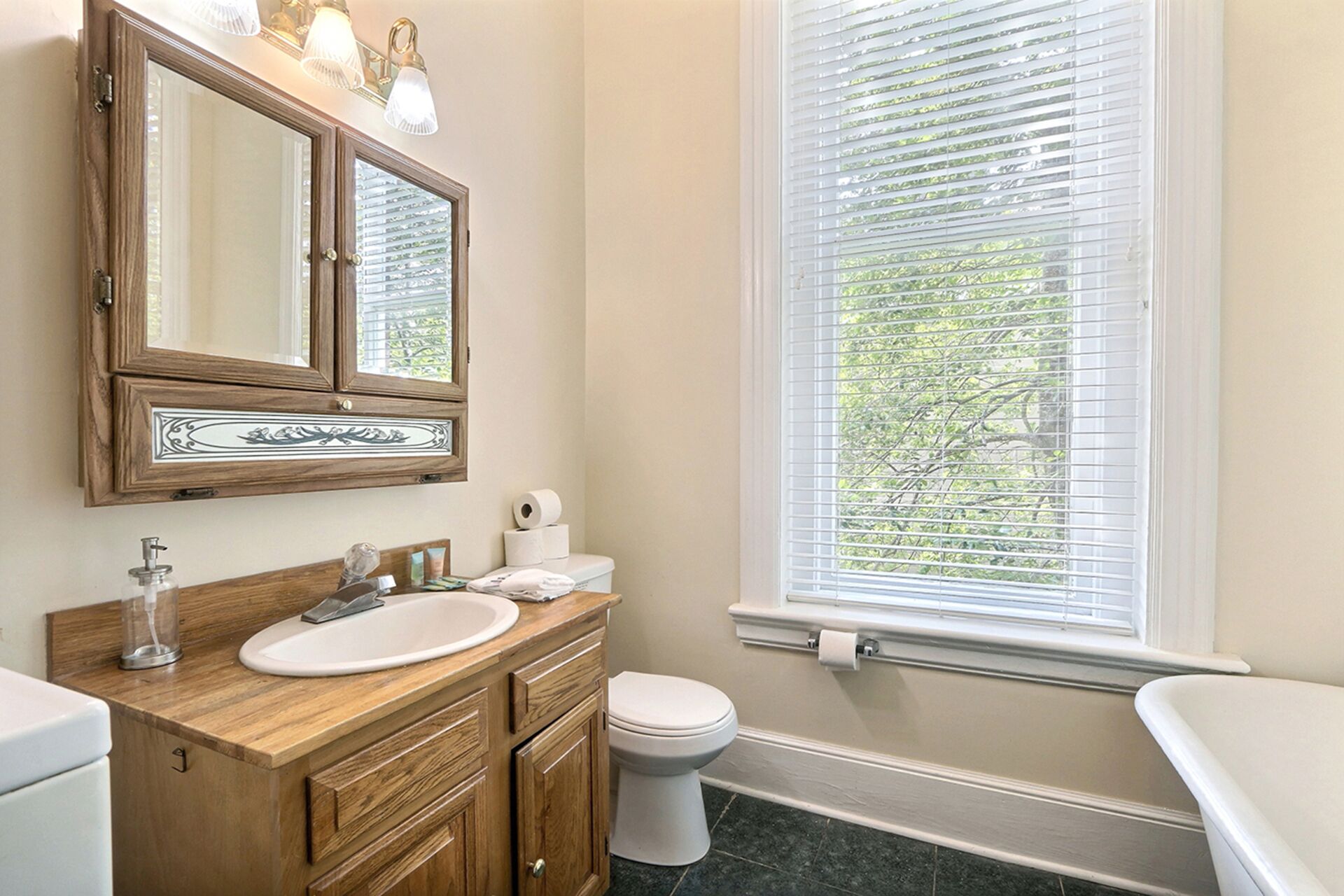 A bathroom with a wooden vanity, a white sink, a toilet, and a window with blinds.