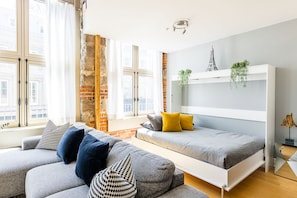 Une chambre moderne avec un lit en mezzanine, un canapé gris agrémenté de coussins à motifs et une vue sur la ville grâce à de grandes fenêtres.