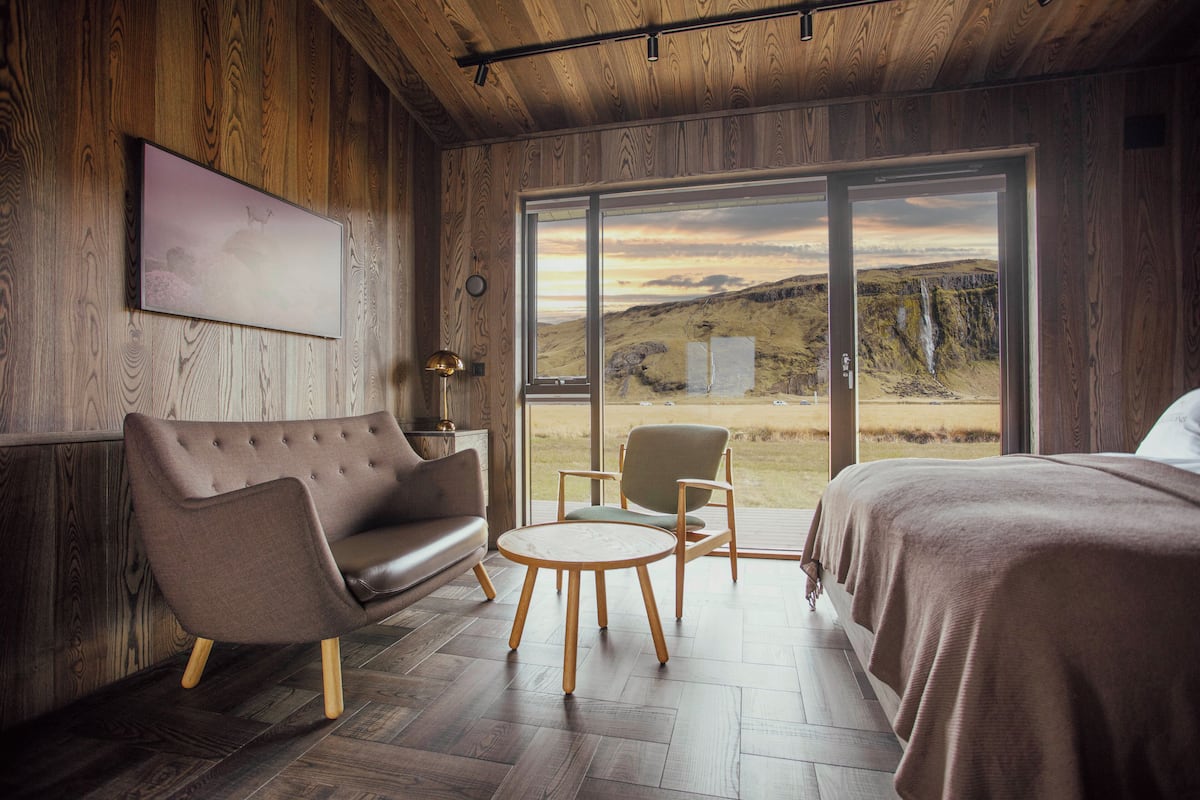 A modern room with a wooden interior, a large window with a view of a landscape, a sofa, a chair, and a bed.