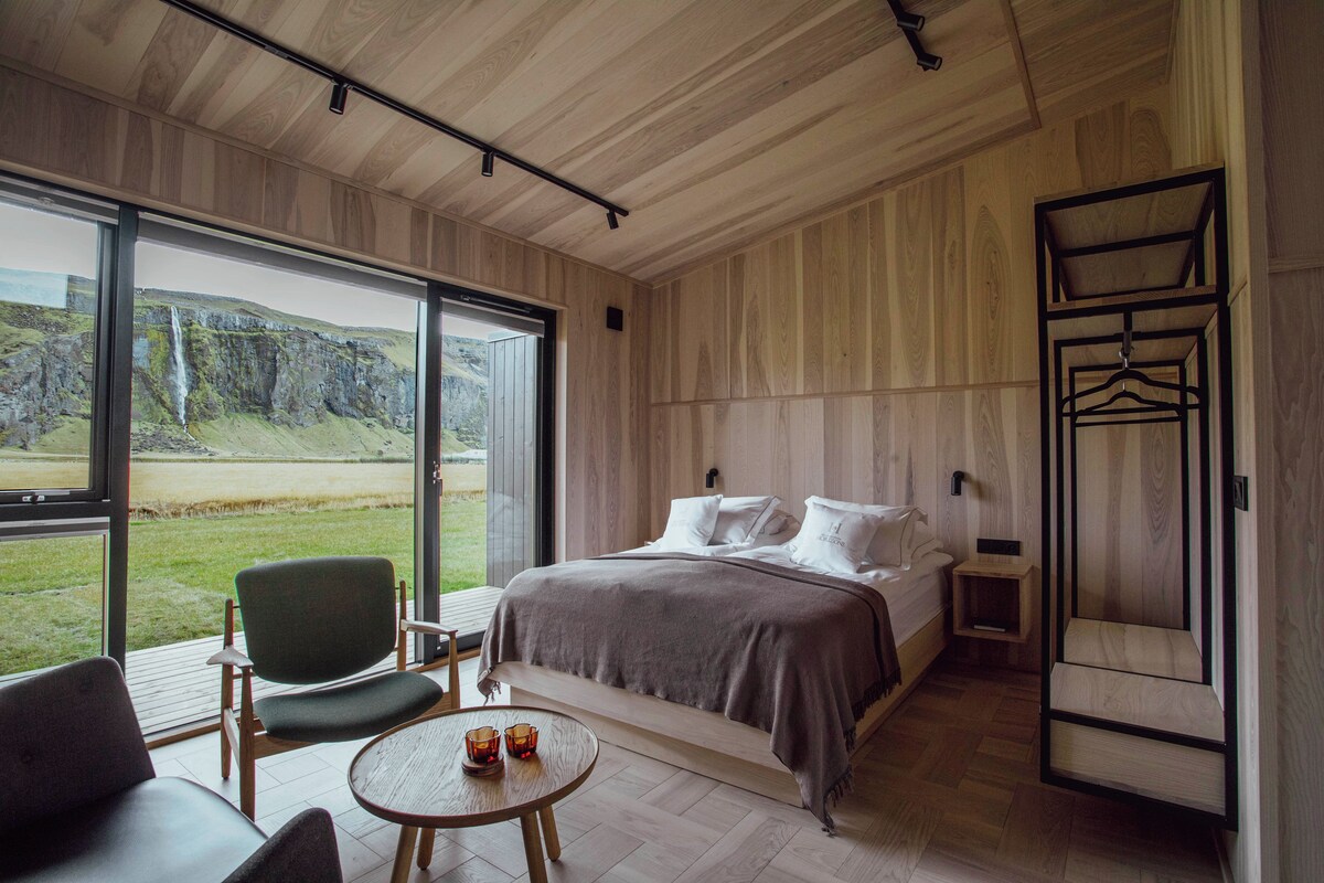 A modern bedroom with a large window, a bed, a chair, a small table, and a wardrobe.