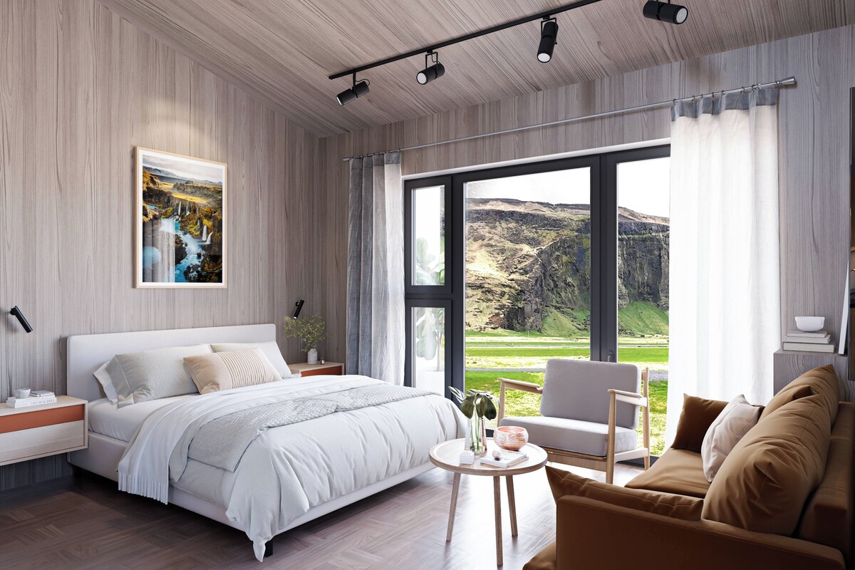 A modern bedroom with a large bed, a bedside table, a nightstand, a round coffee table, a sofa, and a view of a mountainous landscape through large windows.