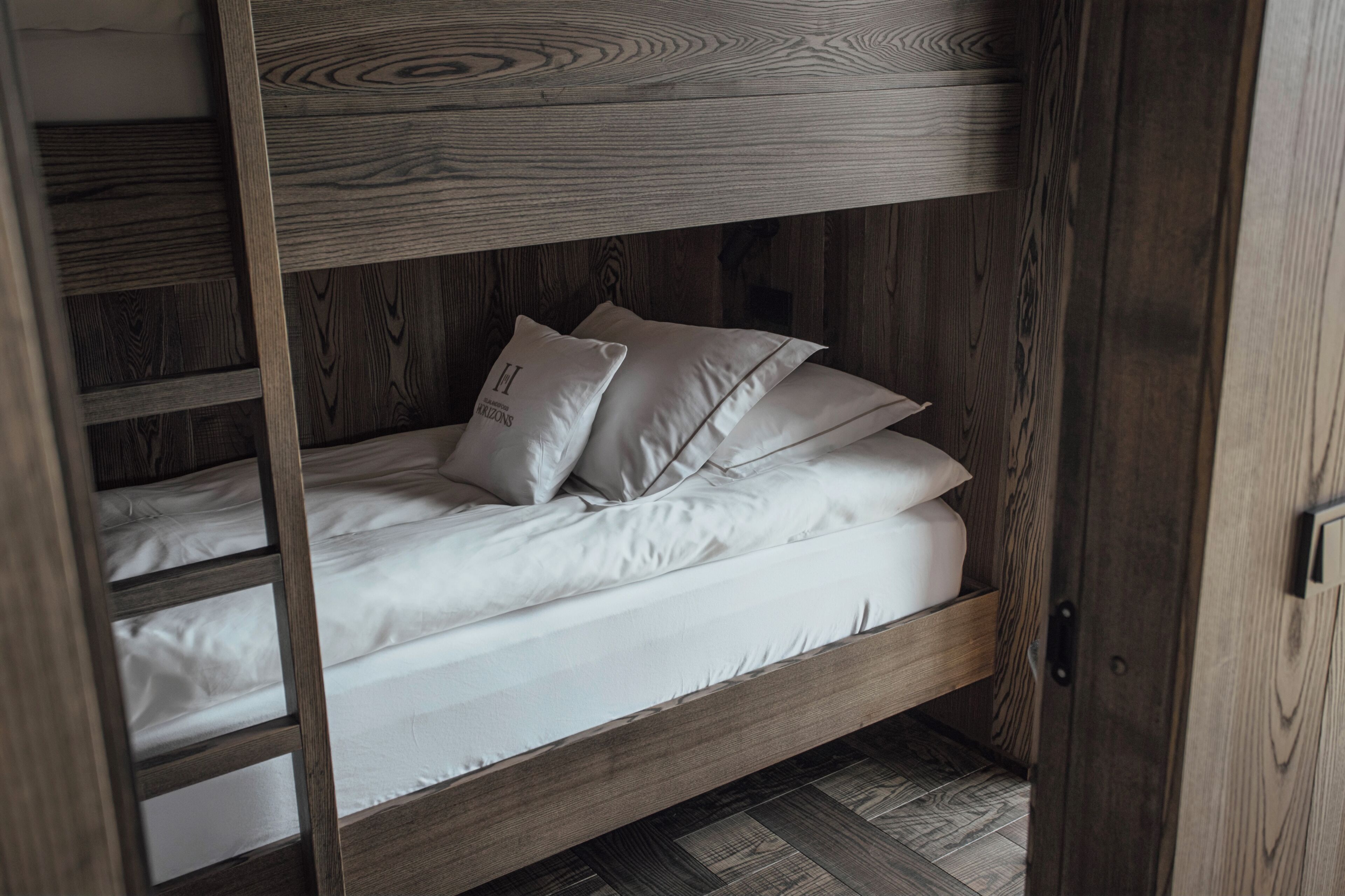 Una cama litera con ropa blanca y almohadas, estructura de madera y una escalera.