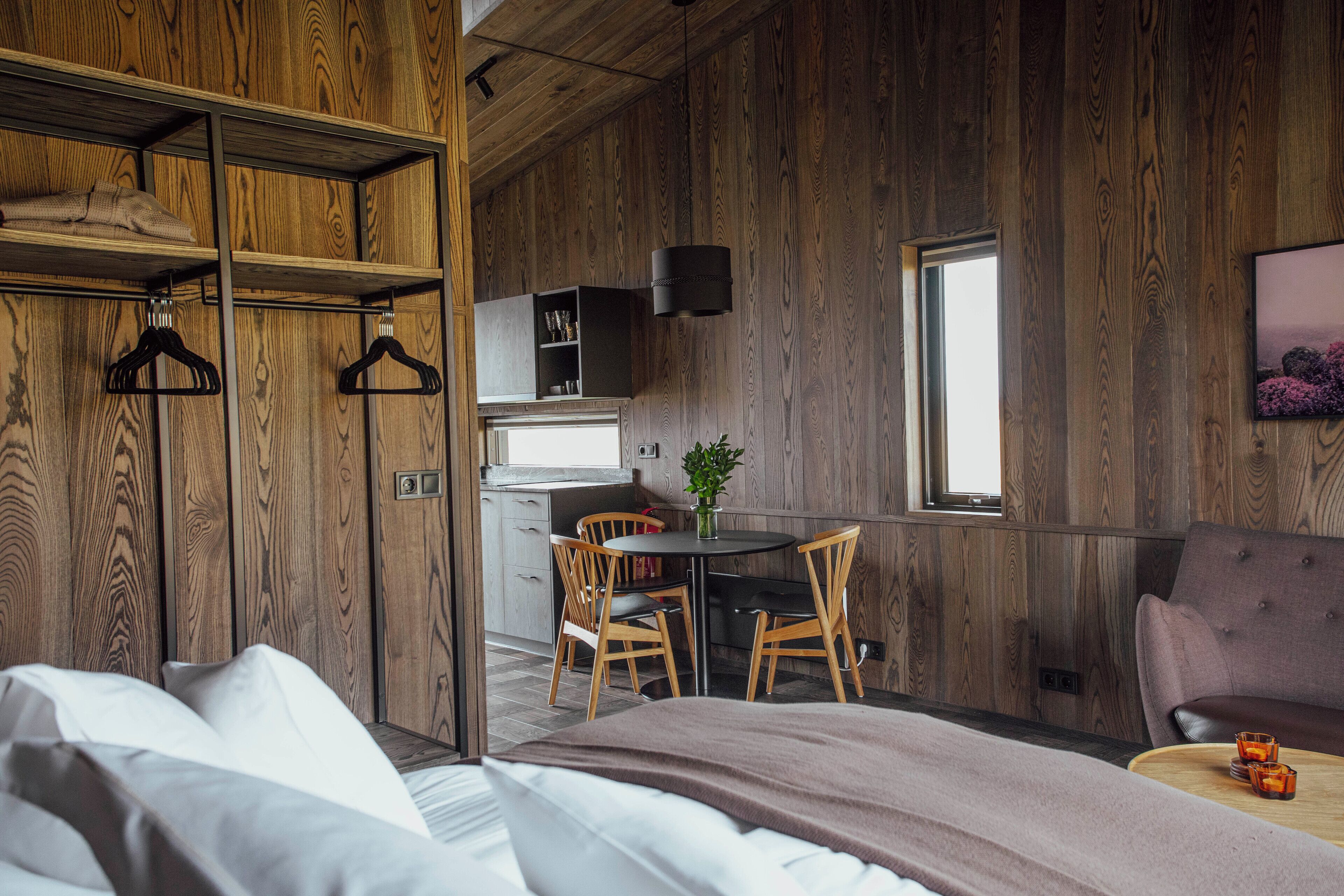 A bedroom with a bed, a wooden wardrobe, a dining area with a table and chairs, a small kitchen, and a window.