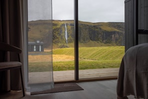 A room with a large window offering a view of a waterfall and a grassy landscape. - Seljalandsfoss Horizons (Rangárþing eystra)
