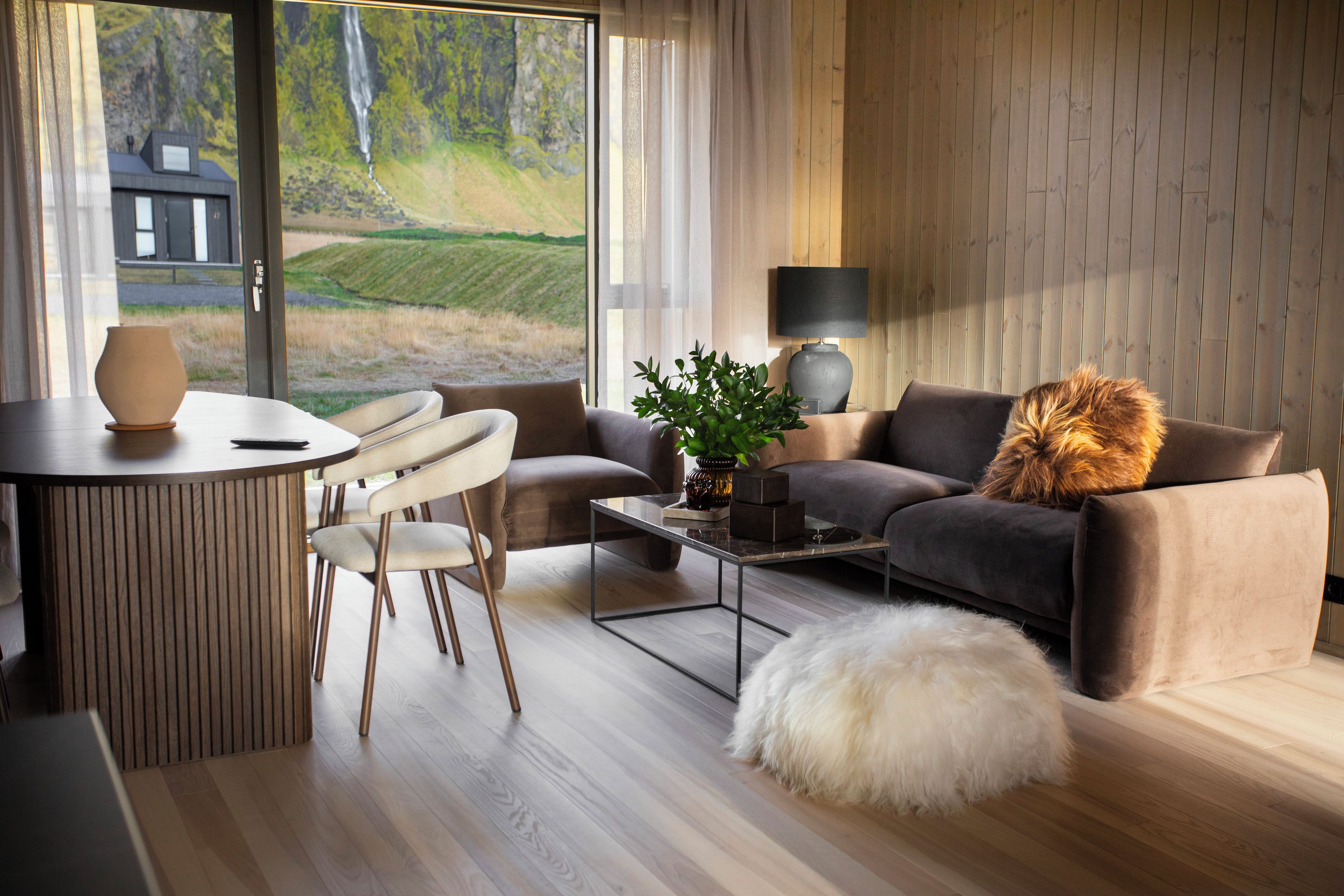 Een moderne woonkamer met een bank, salontafel en stoelen. Grote ramen bieden uitzicht op een huis en een landschap.