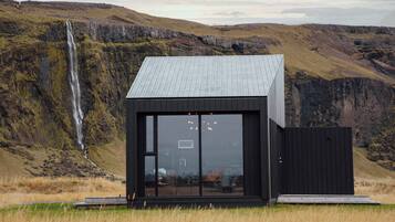 Un edificio moderno, de color negro con grandes ventanales y techo metálico, ubicado en un campo cubierto de hierba con una cascada al fondo.