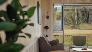 A modern room with a large window overlooking a waterfall. There is a wooden chair and a small table with a lamp. - Seljalandsfoss Horizons (Rangárþing eystra)