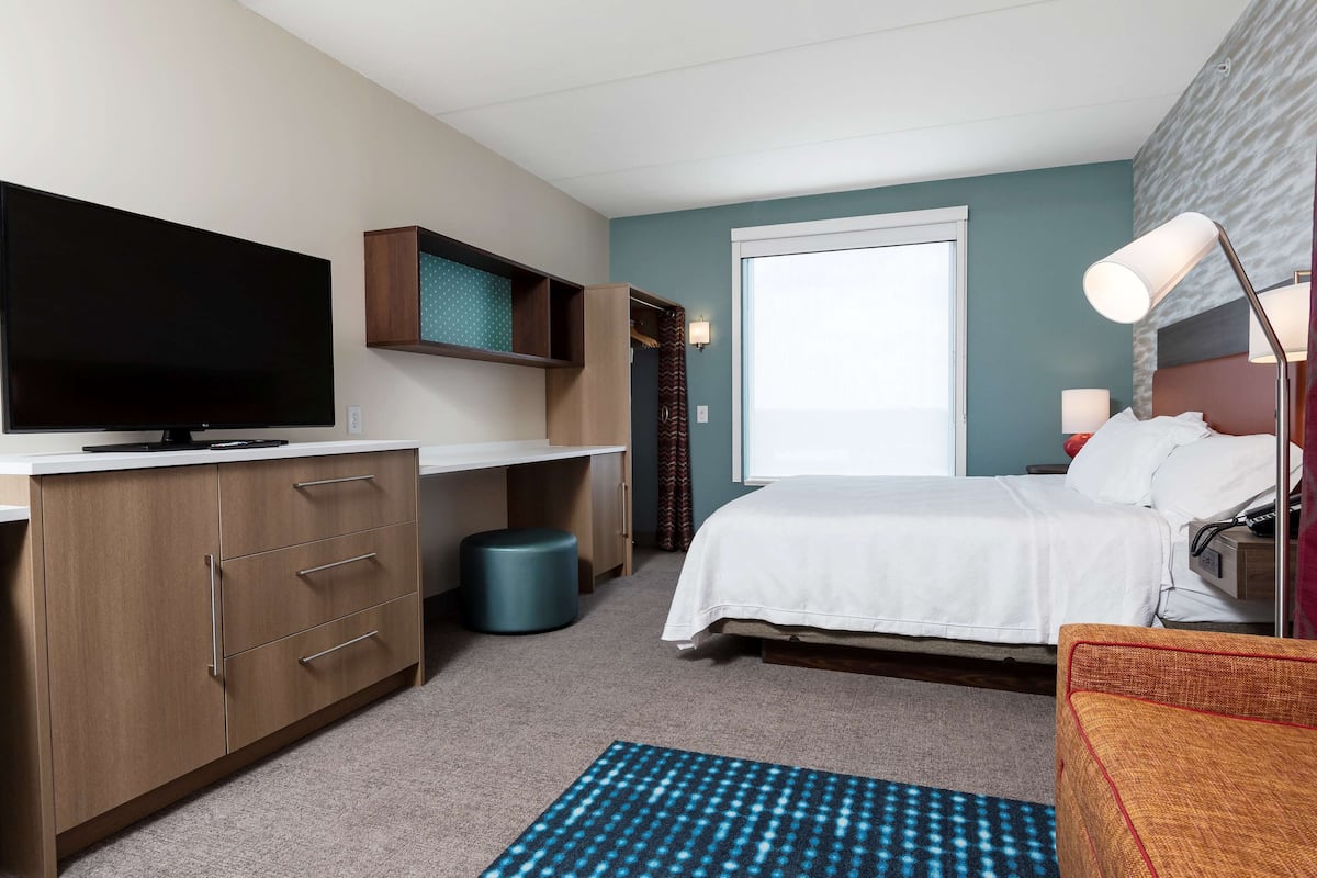 Une chambre d’hôtel équipée d’une télévision à écran plat, d’une armoire en bois, d’un lit avec une literie blanche, d’un tapis à motifs bleus et d’une table de chevet avec une lampe.