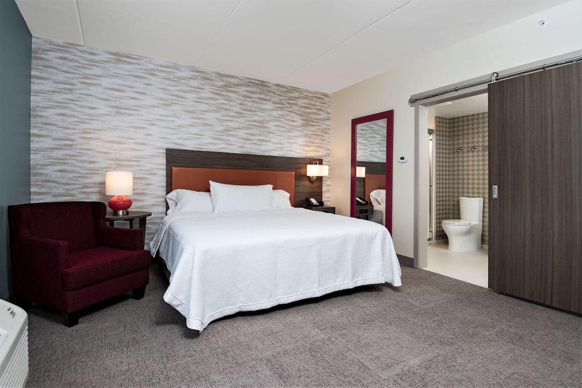 Une chambre d’hôtel avec un grand lit, un fauteuil rouge, une table de chevet avec une lampe, et une salle de bain visible à travers une porte ouverte.