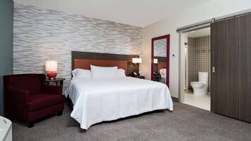 A hotel room with a large bed, a red armchair, a bedside table with a lamp, and a bathroom visible through an open door.