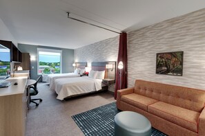 A hotel room with a bed, a desk, a chair, a sofa, and a window with a view of greenery. - Home2 Suites by Hilton Roswell, GA (Alpharetta)